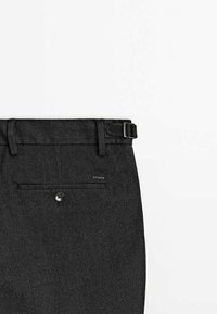 Pantalon de costume gris foncé avec une poche arrière boutonnée et une boucle latérale réglable sur un fond clair uni.