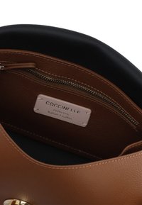 Interno di una borsa in pelle marrone con fodera nera, dotato di una tasca con zip e un'etichetta crema con la scritta "Coccinelle", dal design semplice.