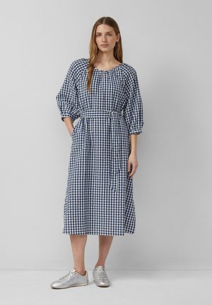 Frau steht mit einer Hand in der Tasche, trägt ein blau-weiß kariertes Midikleid mit Taillengürtel und silbernen Sneakers.