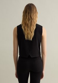 Top nero senza maniche con scollo alto, presenta un design aderente e una fessura sulla schiena. Il modello indossa pantaloni neri abbinati.