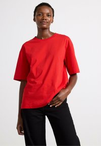 Rode katoenen t-shirt met korte mouwen in een relaxte pasvorm en een ronde halslijn, gecombineerd met zwarte broeken. Gladde textuur en minimalistisch ontwerp.