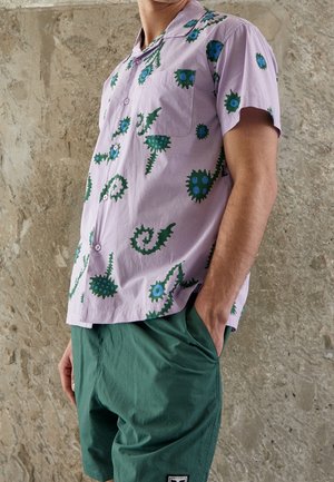 Homme portant une chemise lilas à manches courtes avec des motifs floraux abstraits verts et bleus et un short vert, les mains dans les poches.