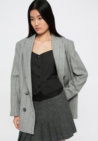 Grå pinstripe blazer over en svart knappetopp, kombinert med et grått plissert skjørt. Stoffet virker teksturert og skreddersydd.