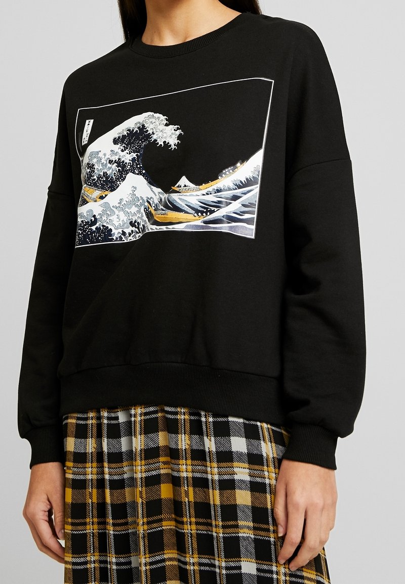 Sweatshirt noir avec un motif de vague blanche agrémenté d'accents bleus et dorés. Poignets et ourlet côtelés. Associé à une jupe à carreaux noire et jaune.