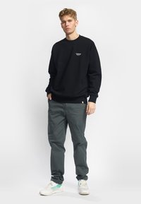 Sudadera negra con cuello redondo y pequeño logo blanco, combinada con pantalones grises y zapatillas de color claro. Un atuendo simple y casual.