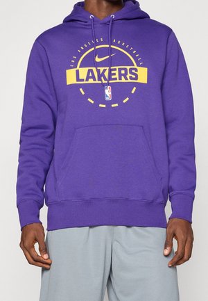 Osoba w fioletowej bluzie z kapturem z żółtymi logo Los Angeles Lakers i Nike oraz szarych szortach, stojąca z rozluźnionymi ramionami wzdłuż boków.