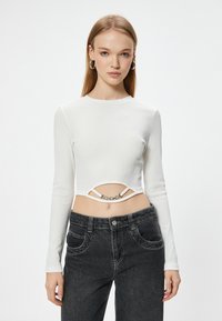 Top corto de manga larga blanco con textura acanalada y un diseño con corte, adornado con una cadena metálica, combinado con jeans negros de cintura alta.