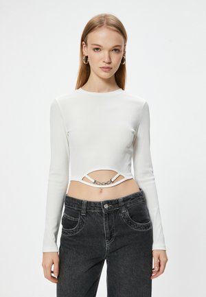 Top corto de manga larga blanco con textura acanalada y un diseño con corte, adornado con una cadena metálica, combinado con jeans negros de cintura alta.