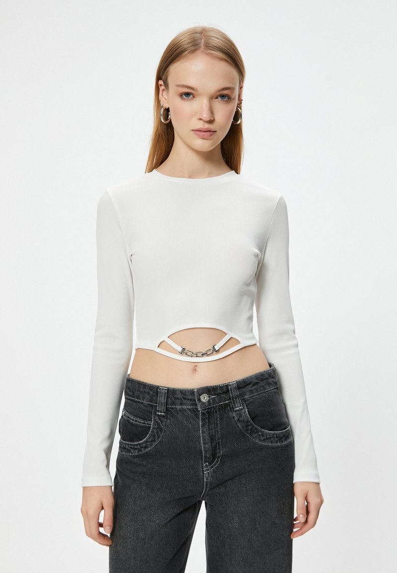 Top corto de manga larga blanco con textura acanalada y un diseño con corte, adornado con una cadena metálica, combinado con jeans negros de cintura alta.