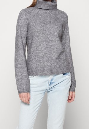 Vrouw draagt een grijze wollen coltrui en lichtblauwe spijkerbroek, staand tegen een effen lichte achtergrond.