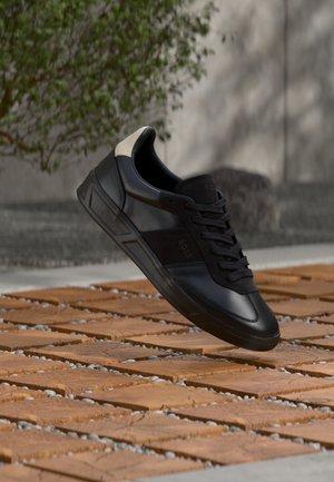 Schwarzer Sneaker in niedriger Form mit Velours- und Lederpaneelen, beigem Fersenakzent und Schnürsenkeln, der über einer gemusterten Holz- und Steinoberfläche im Freien schwebt.