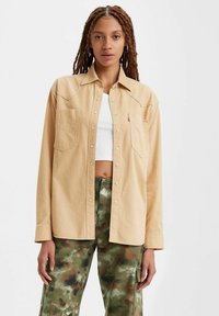 Camicia beige con bottoni e due tasche frontali, dotata di colletto a punta e vestibilità casual, abbinata a pantaloni mimetici verdi.