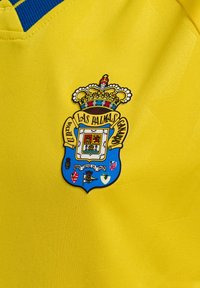 Gul sportsjersey med en blå krave. Inkluderer et broderet våbenskjold med en krone, detaljeret design og forskellige farver.