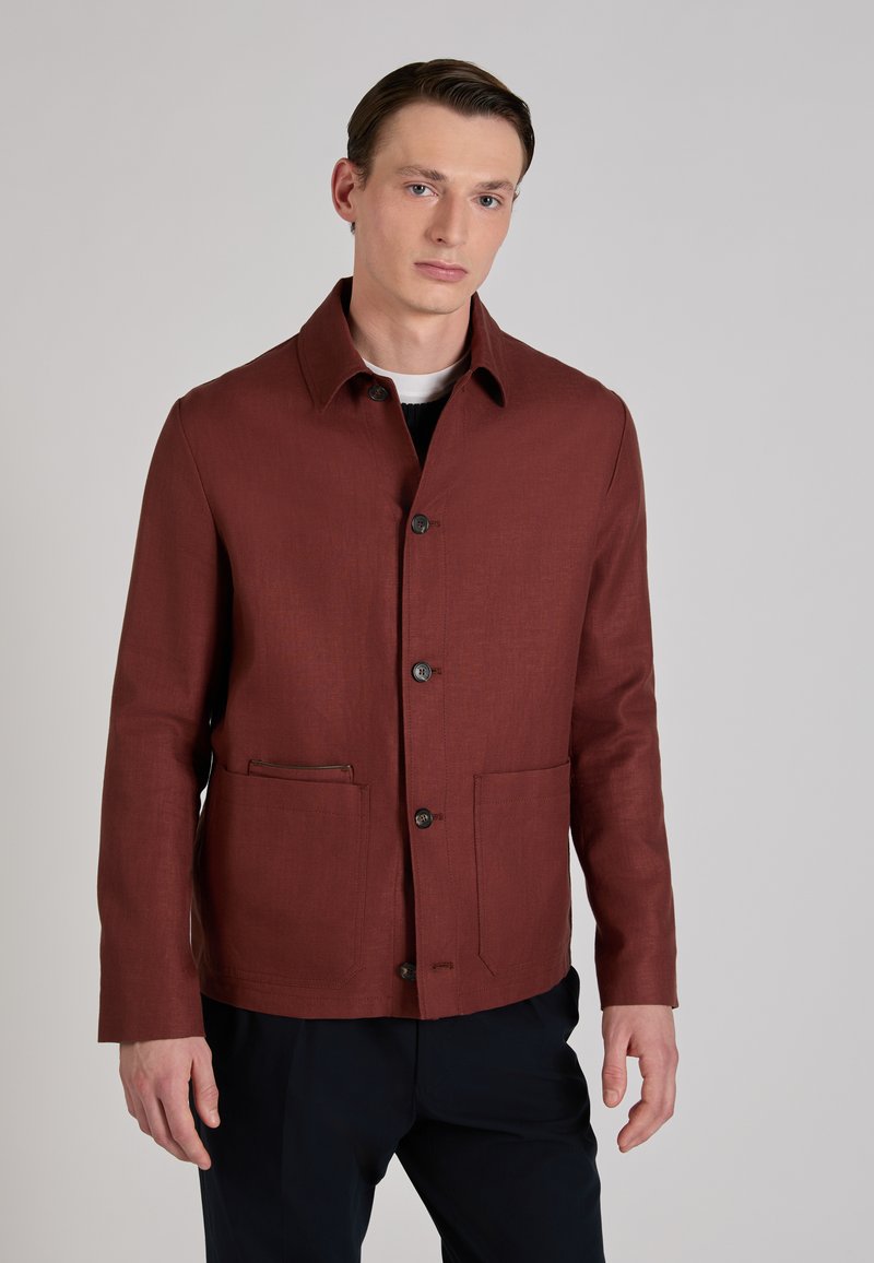 Młody mężczyzna ubrany w zapiętą na guziki rdzawego koloru kurtkę z dużymi przednimi kieszeniami i ciemne spodnie na jednolitym tle.