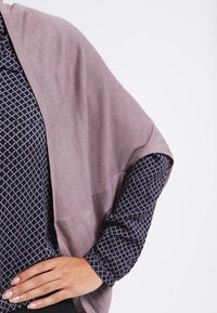 Helle mauve Strickschal mit weicher Textur, drapiert über einer marineblauen gemusterten Bluse. Der Schal hat lose Ärmel und eine lässige Passform.