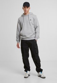 Siv hooded sweatshirt z žepom v obliki kenguruja in črnim logotipom, v kombinaciji s črnimi cargo hlačami z stranskimi žepi, ter belimi supergami z modrimi detajli.