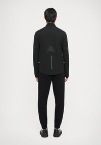 Svart treningsjakke med reflekterende detaljer og prikkete mønster på baksiden, kombinert med svarte joggebukser og teksturerte svarte joggesko.
