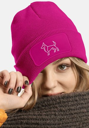 Fuchsia knitted beanie with a ribbed texture and a rectangular patch featuring a white dog graphic. Fingers hold the brim.