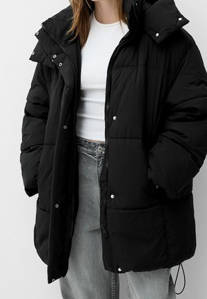 Personne portant une doudoune noire par-dessus un haut blanc ajusté et un jean gris clair, les mains dans les poches de la veste.