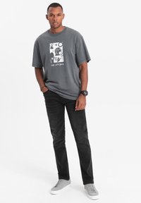 T-shirt in cotone grigio con stampa grafica, vestibilità rilassata, abbinata a jeans neri e sneakers grigie. Design semplice con dettagli minimi.