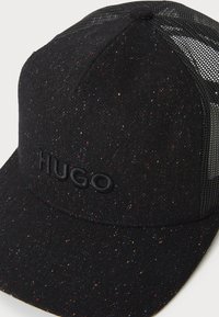 Melnā cepure ar raibām tekstūrām un acs sānu daļām. Piemīt izšuvs "HUGO" logo priekšpusē, nodrošinot strukturētu dizainu.