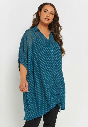 Teal polka dot shirt, sheer fabric, button-up front, collar, short sleeves, asymmetrical hem, soft texture, bold pattern.