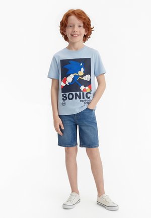 Niño pelirrojo sonriendo, con una camiseta gráfica de Sonic the Hedgehog de color azul claro, pantalones cortos de mezclilla y zapatillas blancas sobre un fondo blanco.