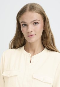 Cream-colored blouse with a mandarin collar, two front pockets, and button closures at the neck. Smooth texture and lightweight material.