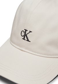 Gorra beige hecha de tela texturizada, con un logotipo bordado en negro "CK" en la parte frontal y costuras visibles a lo largo de la visera.