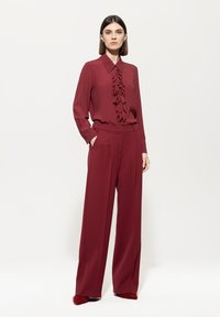 Burgundy blouse with ruffled front detailing, paired with loose-fitting trousers. The outfit features a smooth texture and tailored design.