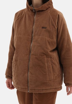 Persona que lleva una chaqueta Vans de pana marrón con cremallera, puños elásticos y bolsillos laterales, mostrada desde los hombros hasta la mitad del muslo.