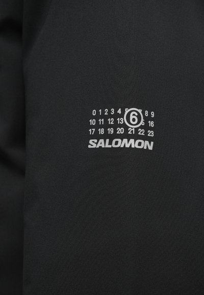 Fekete szövet puha textúrával, amelyen egy fehér "SALOMON" logó és egy számozott minta található, ami az órákat 0-tól 23-ig jelöli, a 6-os szám körül pedig egy kör van.