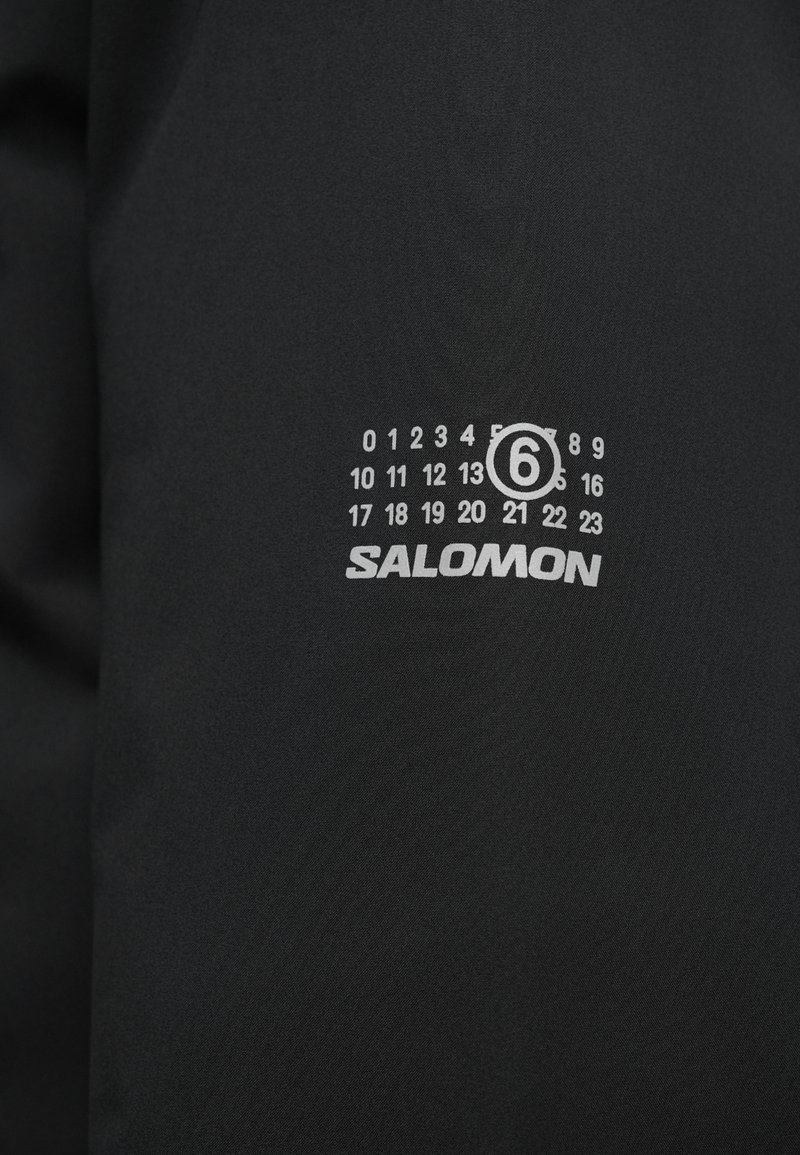 Sort stof med en blød tekstur prydes af et hvidt logo "SALOMON" og et nummereret design, der angiver timer fra 0 til 23, med en cirkel omkring tallet 6.