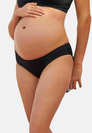 Donna incinta che indossa biancheria intima nera e reggiseno, toccandosi la pancia con una mano e tenendo gli occhiali da sole con l'altra.
