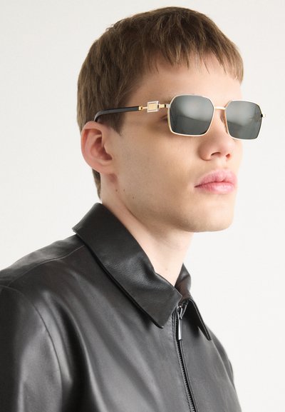 Gafas de sol con montura octagonal, detalles en metal dorado, lentes grises y una vara negra. Modelo vistiendo una chaqueta de cuero negra.