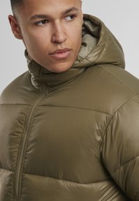 Olíva zöld puffernyakú dzseki magas gallérral és kapucnival, sima, fényes textúrával és láthatóan varrott részletekkel az illesztések mentén.