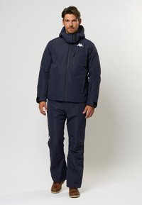 Mörkblå isolerad skidjacka med hög krage, dragkedja och logotyp; matchande byxor med rak skärning; slitstarkt tyg; slät textur.