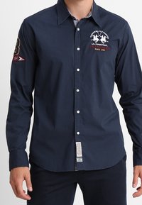 Camisa de manga larga azul marino con botones, fabricada en algodón. Cuenta con un logo en el pecho izquierdo y un parche con el número 3 en la manga derecha.