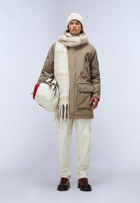 Khaki pufferjakke med frontlommer, cremet tørklæde med striber, cremebukser og brune støvler med røde snørebånd, holder en beige taske.