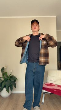 Raiba brūna un bēša flīsa jaka virs melna T-krekla, savienota ar ziliem denim džinsiem un gaiši pelēkiem slip-on apaviem. Telpu vide.