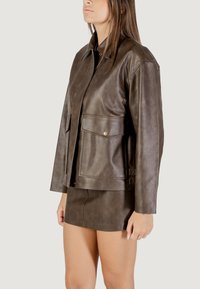 Giacca in finta pelle marrone con colletto classico, due tasche sul petto con chiusura a bottone e vestibilità rilassata, abbinata a una gonna coordinata.