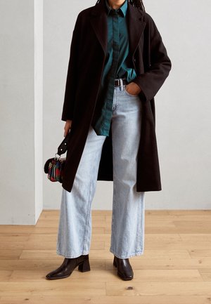 Personne portant un manteau marron foncé, une chemise sarcelle, un jean large bleu clair et des bottines foncées, tenant un sac à motifs multicolores.