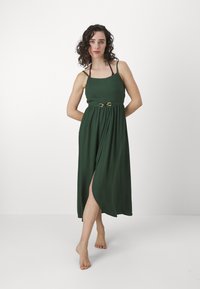 Vestido maxi verde com um corpete ajustado, alças finas e uma cintura cingida. O tecido cai elegantemente, apresentando uma fenda frontal e uma textura suave.