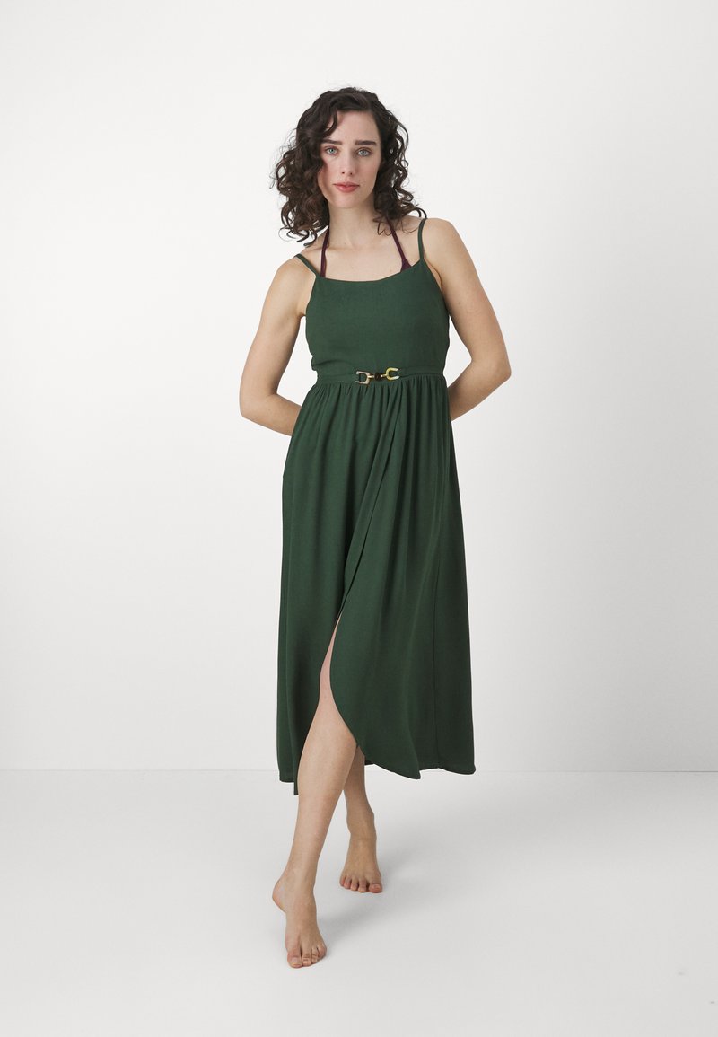 Vestido maxi verde com um corpete ajustado, alças finas e uma cintura cingida. O tecido cai elegantemente, apresentando uma fenda frontal e uma textura suave.