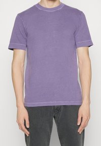 Fioletowy krótki rękaw T-shirt wykonany z miękkiej bawełny, z okrągłym dekoltem i widocznymi detalami przeszycia. Noszony z ciemnoszarymi spodniami.