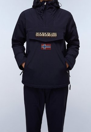 Personne portant une veste à capuche Napapijri bleu marine, les mains dans les poches, avec un écusson du drapeau norvégien au centre avant.