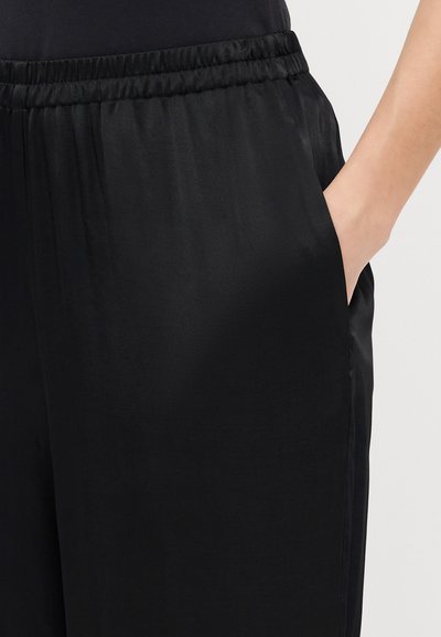 Svarta satinkjolbyxor med elastisk midja, sidofickor och en slät yta, delvis visade med en hand i en ficka.