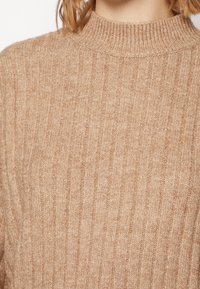 Beżowy prążkowany sweter z wysokim dekoltem. Miękka faktura o luźnym kroju, z pionowymi paskami na całej tkaninie.