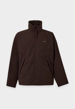 Homme portant une veste marron foncé, un col roulé bleu marine, un pantalon marron large et des chaussures foncées et brillantes, posant devant un fond blanc uni.