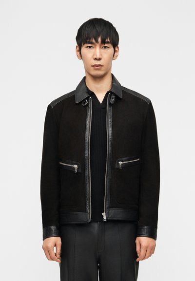Homme portant une veste noire avec finitions en cuir et poches zippées par-dessus un pull à col en V côtelé noir, debout devant un fond blanc uni.